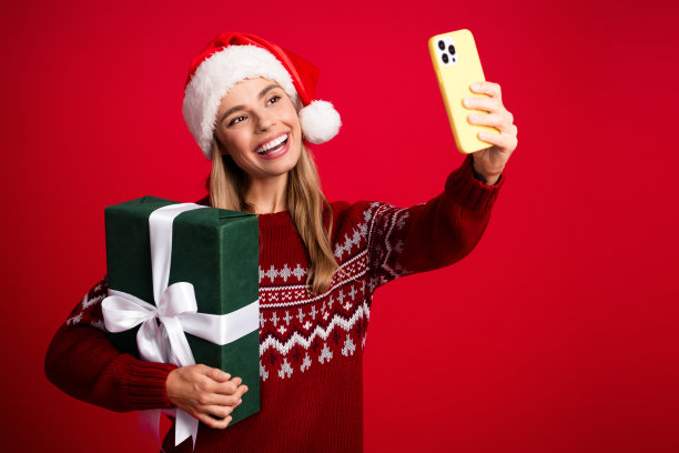 戴着圣诞帽、穿着红色毛衣的俏皮女子，手持一个大大的绿色礼物，在鲜艳的红色背景前开心地自拍，用于圣诞促销图库照片图片下载