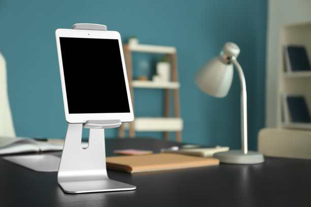 一张照片，展示了一台通用的数字平板电脑，屏幕空白，竖直放置在一个金属支架上，支架放在深色的办公桌上。图片下载