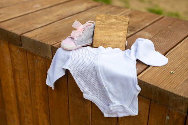 木质表面上放着婴儿连体衣、粉色和灰色鞋子，以及一个暗示新生儿即将到来的小木牌。木牌上留有写下宝宝出生日期的位置。图片下载