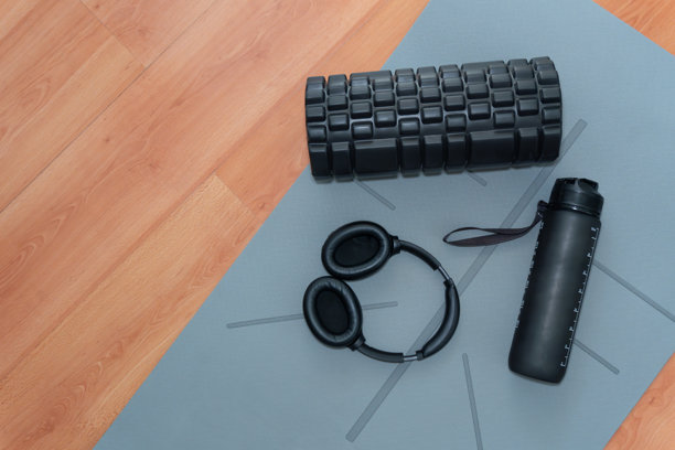 从上方拍摄的家庭健身必需品照片：黑色的泡沫轴、耳机和水瓶，放在木地板上的灰色瑜伽垫上。图片下载