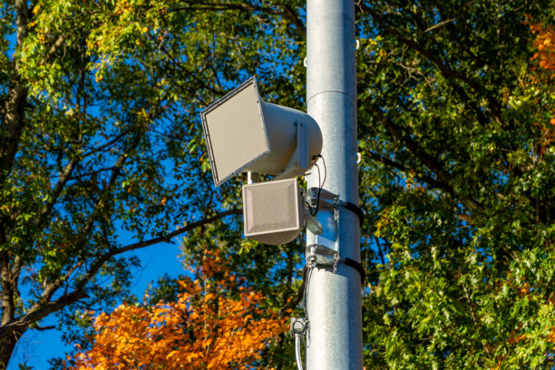 一张体育赛事中常见的户外体育场扬声器的照片，安装在镀锌灯杆上，背景有树木。图片下载