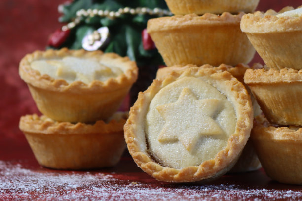 全画幅图片，一个单独的肉馅饼靠在一堆肉馅饼上，顶部有裹着糖粉的酥皮星星盖子，一叠节日甜点酥皮饼，圣诞树模型，斑驳的红色背景，焦点在前景。图片下载