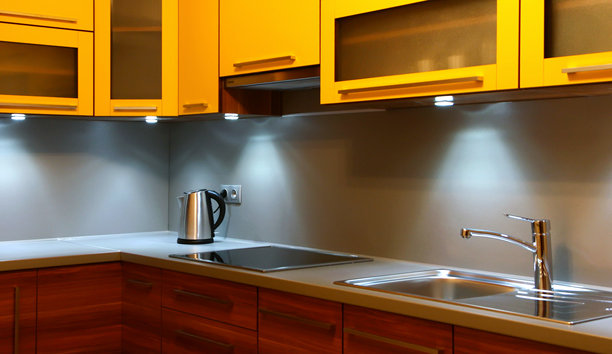 带橱柜灯照明的现代厨房内部，配有黄色橱柜、木制抽屉、不锈钢水槽、水龙头和电热水壶图片下载