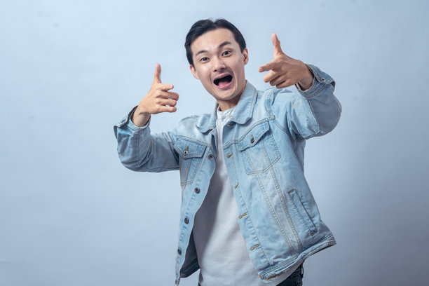 身穿牛仔夹克的亚洲男子，自信地微笑，比出“枪手”手势，背景简洁，概念用于成功、积极、自我表达和现代生活方式内容图片下载