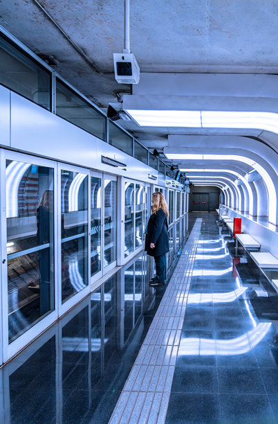 一张在巴塞罗那Hospitalet的Ciutat de la Justícia地铁站，一位孤独、沉思的女性在蓝色灯光的未来感地铁站台上等待列车的竖幅照片。图片下载