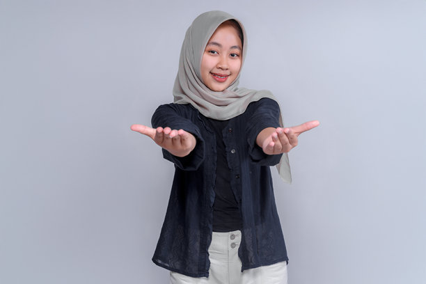 一位年轻的亚洲女性，戴着头巾，身穿黑色衬衫，面带微笑，伸出手做出欢迎的姿势。这张照片传达出友好、温暖和积极的氛围，非常适合商业用途。图片下载
