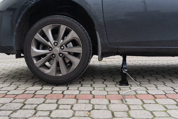 一张近距离的户外照片，展示了一辆深色轿车被一个机械剪刀千斤顶抬起，停放在铺有格子方砖的车道上，准备更换轮胎。图片下载