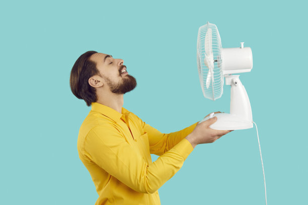 一个留着胡子的男人，在浅蓝色背景下，用电风扇吹风，享受新鲜空气，躲避炎热。图片下载