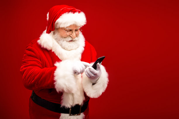 圣诞老人使用智能手机在明亮的红色工作室摆姿势，拍摄圣诞节主题的素材图片图片下载