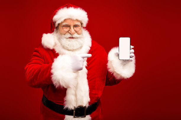 圣诞老人举着空白的智能手机屏幕，在明亮的红色背景前指向它，用于圣诞主题的素材图片图片下载