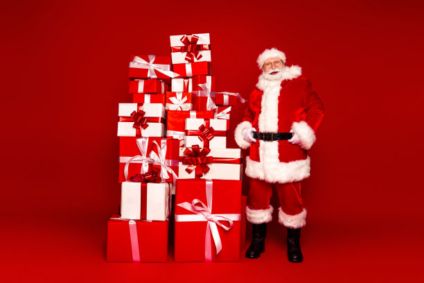 身穿经典圣诞老人服装的圣诞老人站在一堆高高的礼物盒旁，为节日拍摄照片图片下载