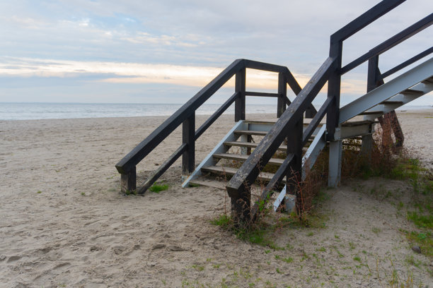 一张风化的木制楼梯照片，黑色栏杆通向宽阔宁静的沙滩，沙滩延伸至海天相接的平静海面，天空多云。图片下载