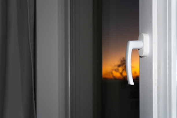 特写照片，展示了一个白色的PVC窗户把手，已旋转至开启或通风位置，背景为黑暗的室内和戏剧性色彩的户外日落景象。图片下载
