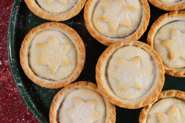 一张近景图片，画面为绿色盘子盛放的肉馅饼，上面覆盖着星星形状的酥皮盖，这是节日圣诞节的甜点，酥皮点心内填有甜肉馅，撒有糖粉，背景为斑驳的红色，俯视角度。图片下载