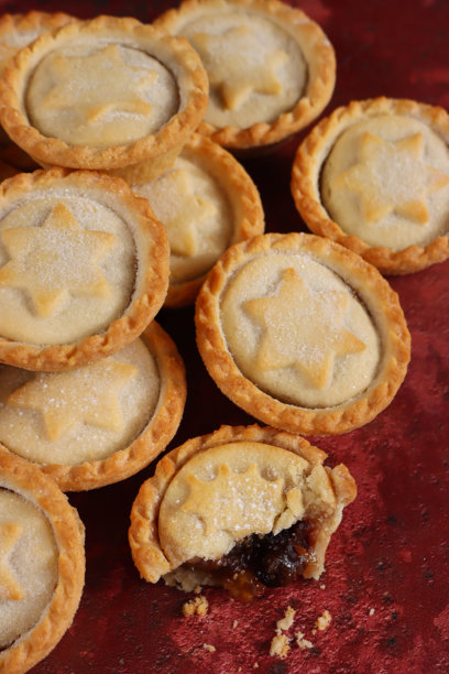 特写图片，展示堆叠的肉馅饼，上面覆盖着带有糕点星星和冬青叶的精美盖子，节日圣诞甜点酥皮糕点横截面展示甜肉馅，撒上糖粉，斑驳的红色背景，俯视角度图片下载