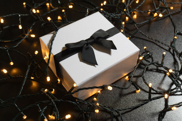 特写照片，一个优雅的白色礼品盒，用有纹理的黑色丝带系着，周围环绕着发光的、闪烁的串灯。图片下载