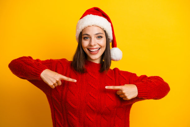 戴着圣诞帽的年轻女子穿着红色毛衣，指着自己，面带笑容，拍摄节日主题照片图片下载