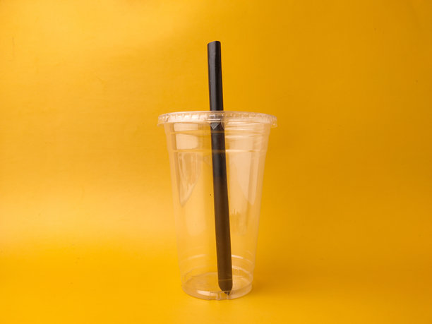 图片展示了一个透明的一次性塑料杯，配有杯盖和黑色吸管，背景为黄色。图片下载