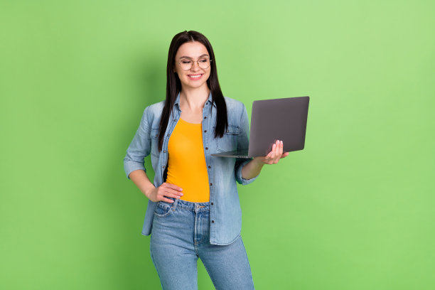一位快乐友善的棕色头发女士戴着眼镜，穿着牛仔裤和衬衫，橙色上衣，孤立在绿色背景上，正在看笔记本电脑的照片图片下载