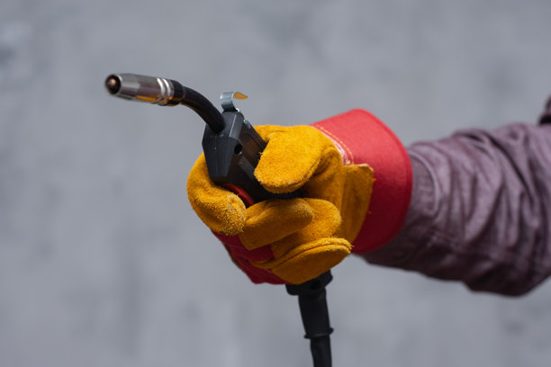 戴着红黄相间防护手套的工人的手部特写，手中握着焊枪。该图片代表了工业劳动、焊接过程和安全设备。图片下载