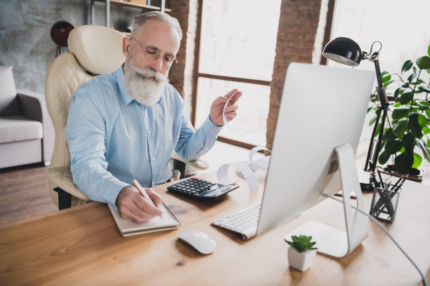 照片：严肃专注的老年退休会计在办公室工作站内写笔记图片下载