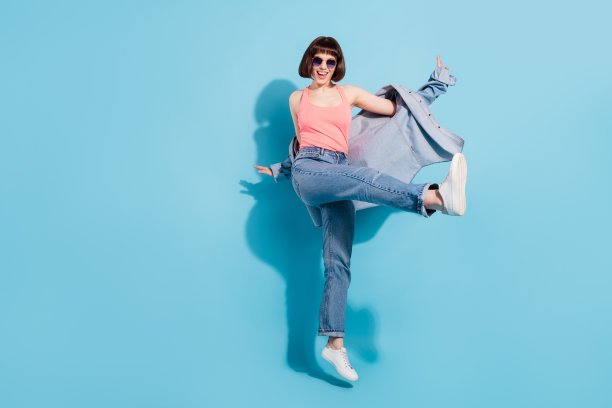 身穿粉色上衣、牛仔裤和眼镜的快乐年轻女子，在蓝色背景前尽情欢呼舞蹈的全身照片图片下载