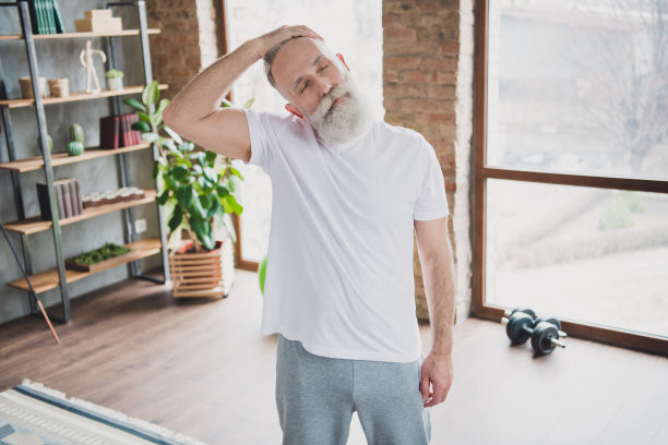 照片：一位心情愉悦、充满活力的爷爷在家中锻炼，伸展脖子，享受居家训练的时光。图片下载