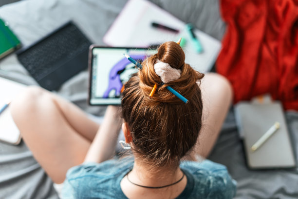 女孩盘腿坐在床上，俯视视角，手持触控笔和数位板，正在绘制色彩鲜艳的卡通插画。周围摆放着创作工具，营造出创意随性的氛围。图片下载