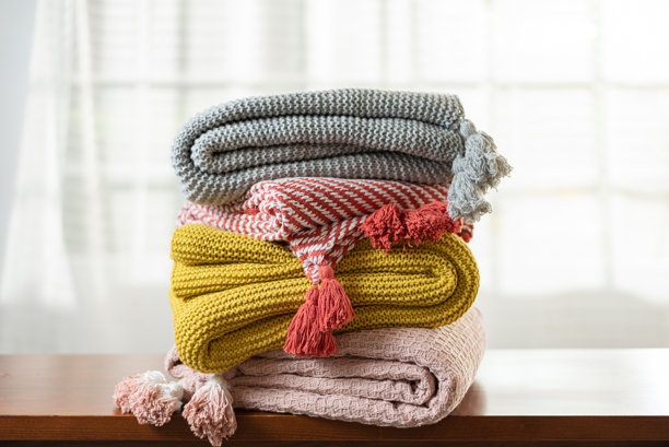 一张照片，展示了四张色彩鲜艳、纹理独特的针织毯子整齐地堆叠在一起，上面装饰着流苏，颜色分别为灰色、芥末黄、红白条纹和灰粉色，放置在木质表面上。图片下载