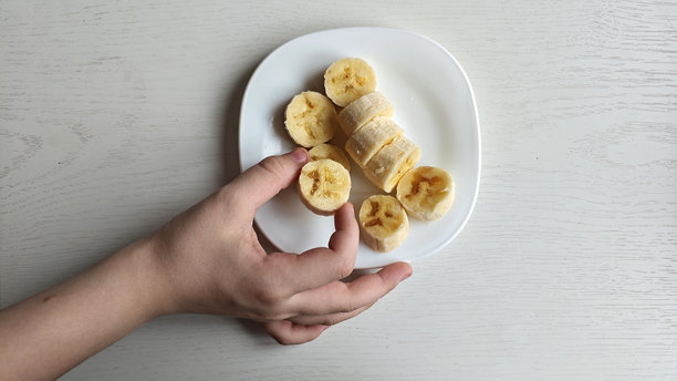 一只手拿着香蕉片，放在一个白色盘子上方，盘子上已有几片香蕉。盘子放置在浅色木质表面上。香蕉片在盘子上呈圆形排列。健康饮食。高质量照片。图片下载