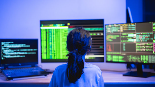 在昏暗的环境中，一个年轻人正在与多个电脑屏幕互动，屏幕上显示着代码和数据分析，并伴有蓝色灯光效果。图片下载