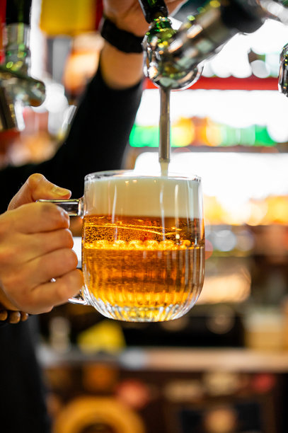 酒保的手在酒吧或酒馆环境中，将新鲜的金黄色啤酒从酒龙头倒入冰镇的、带肋的玻璃马克杯中，杯中充满了浓密、绵密的泡沫。图片下载