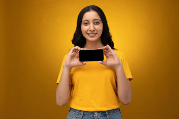 年轻女子展示手机屏幕，背景为黄色隔离的素材照片图片下载