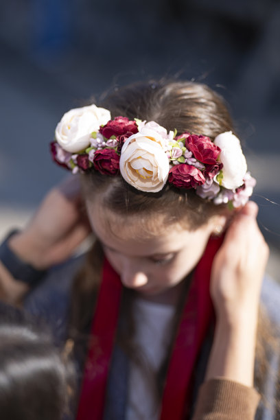 特写竖幅照片，一位母亲拥抱着她悲伤的女儿，女儿戴着花环，脖子上围着灰色夹克，焦点清晰，寓意着爱与情感支持。图片下载