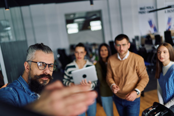 一群年轻的专业人士聚集在现代办公室进行商务演示。一位留着胡须的男士正在大屏幕上讲解数据图表，团队成员则专注倾听。体现团队合作、领导力以及数据驱动策略的概念。图片下载