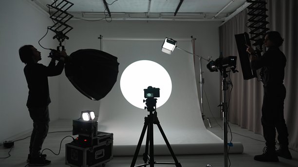 摄影师和助理正在设置灯光和相机，在带有现代照明设备和大型白色背景幕的专业摄影棚进行照片和视频制作拍摄。图片下载
