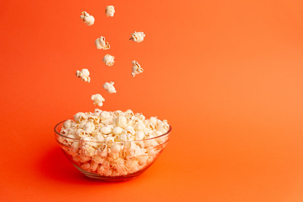 橙色背景下，爆米花掉入玻璃碗的垂直照片，充满动感的场景，非常适合用于食物、电影或生活方式概念。图片下载