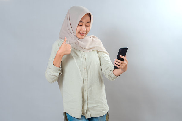 快乐的年轻穆斯林女性戴着头巾，竖起大拇指，同时看着智能手机。她面带微笑，表情积极，这是一张用于社交媒体或广告的垂直工作室照片。图片下载