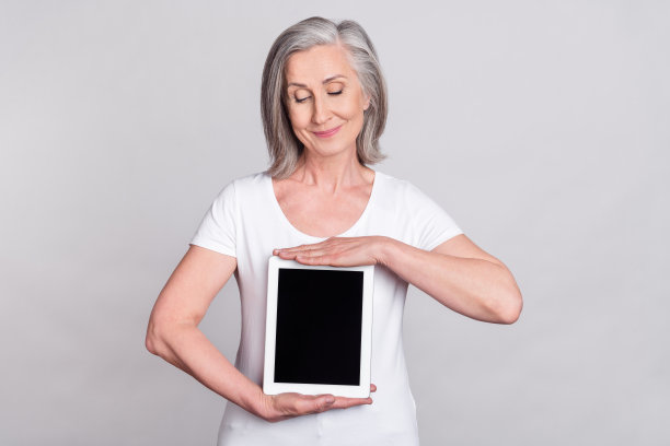 照片：漂亮的、快乐的、年长女经理穿着白色T恤，手持平板电脑，孤立在灰色背景上。图片下载