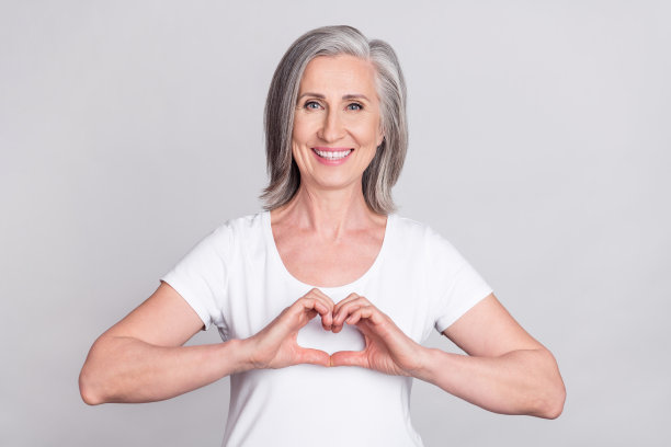 照片：一位年长的女性带着快乐积极的微笑，比出手指爱心符号，在灰色背景上展示爱意，背景为灰色。图片下载