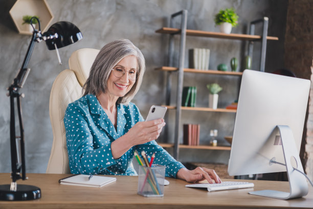 照片：一位面带微笑、头发灰白、快乐积极的老年女性，坐在办公室工作区的桌子旁，看着手机图片下载