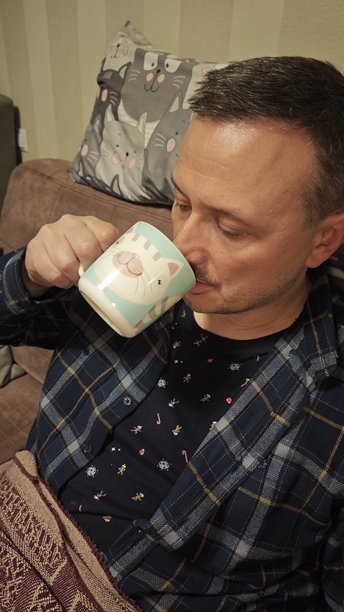 一个男人在家中舒适的氛围里，惬意地坐在沙发上，喝着印有可爱猫咪图案的马克杯里的茶。这张图片象征着舒适、放松和夜晚的自我关怀。图片下载