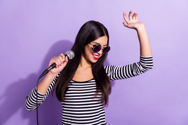 身着酷炫太阳镜、心情愉悦的中国女孩，正在紫色背景前开心唱卡拉OK、跳舞的照片图片下载