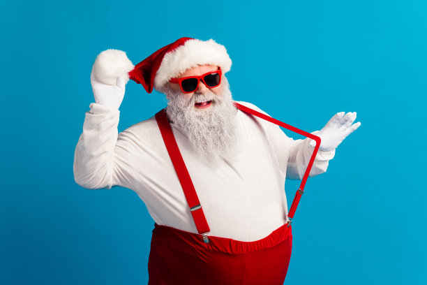 穿着红色吊带和帽子的俏皮圣诞老人，在蓝色影棚里微笑，捕捉圣诞节的魔力与节日欢乐的素材照图片下载