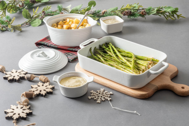 照片展示了白色的陶瓷餐具，里面盛满了烤芦笋和土豆，放在一块砧板上，准备迎接圣诞晚餐，周围环绕着节日常青树和雪花装饰。图片下载