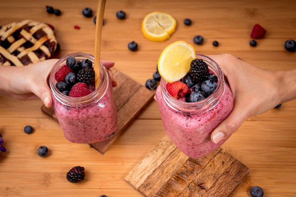 俯视拍摄的特写照片，展示了两个人手中拿着装有健康粉色浆果冰沙的小玻璃罐，冰沙顶部饰有新鲜覆盆子、黑莓和柠檬片。图片下载