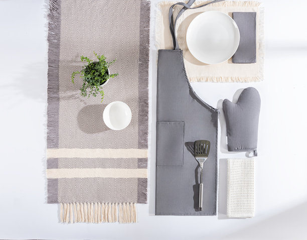 白色表面上整齐摆放着各种灰色和米色厨房纺织品及烹饪配件的照片，包括围裙、烤箱手套、桌布和餐具。图片下载