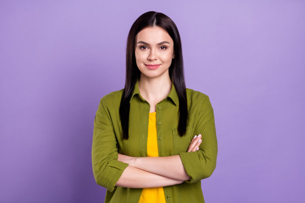 身穿绿色服装、双臂交叉、面带微笑的时尚迷人年轻女子照片，背景为紫色图片下载