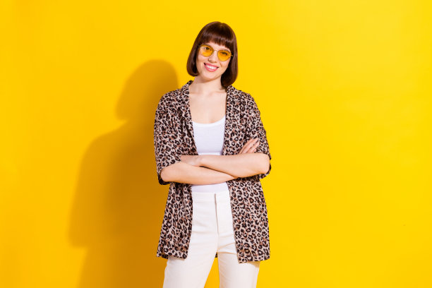 照片：一位乐观的年轻女子，双臂交叉，戴着眼镜，穿着豹纹衬衫，孤立在黄色背景上。图片下载