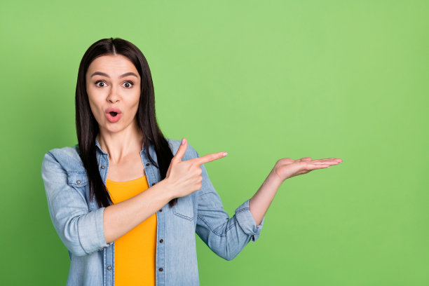 照片：留着棕色长发的可爱女士，身穿牛仔衬衫，表情惊讶，手指指向空白处，背景为淡绿色图片下载